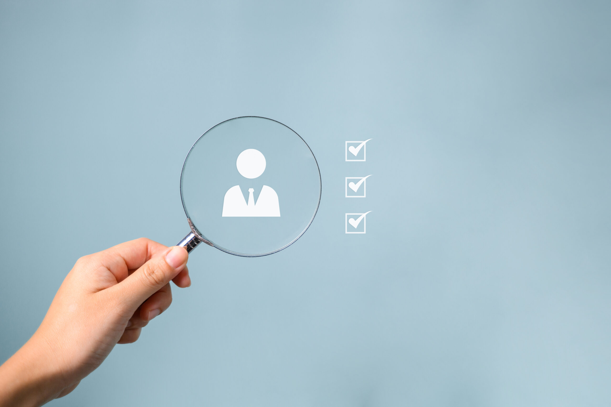 Hand holding a magnifying glass over an employee icon with checklist marks, symbolizing HR overload, compliance uncertainty, turnover issues, and the need to outsource HR support.